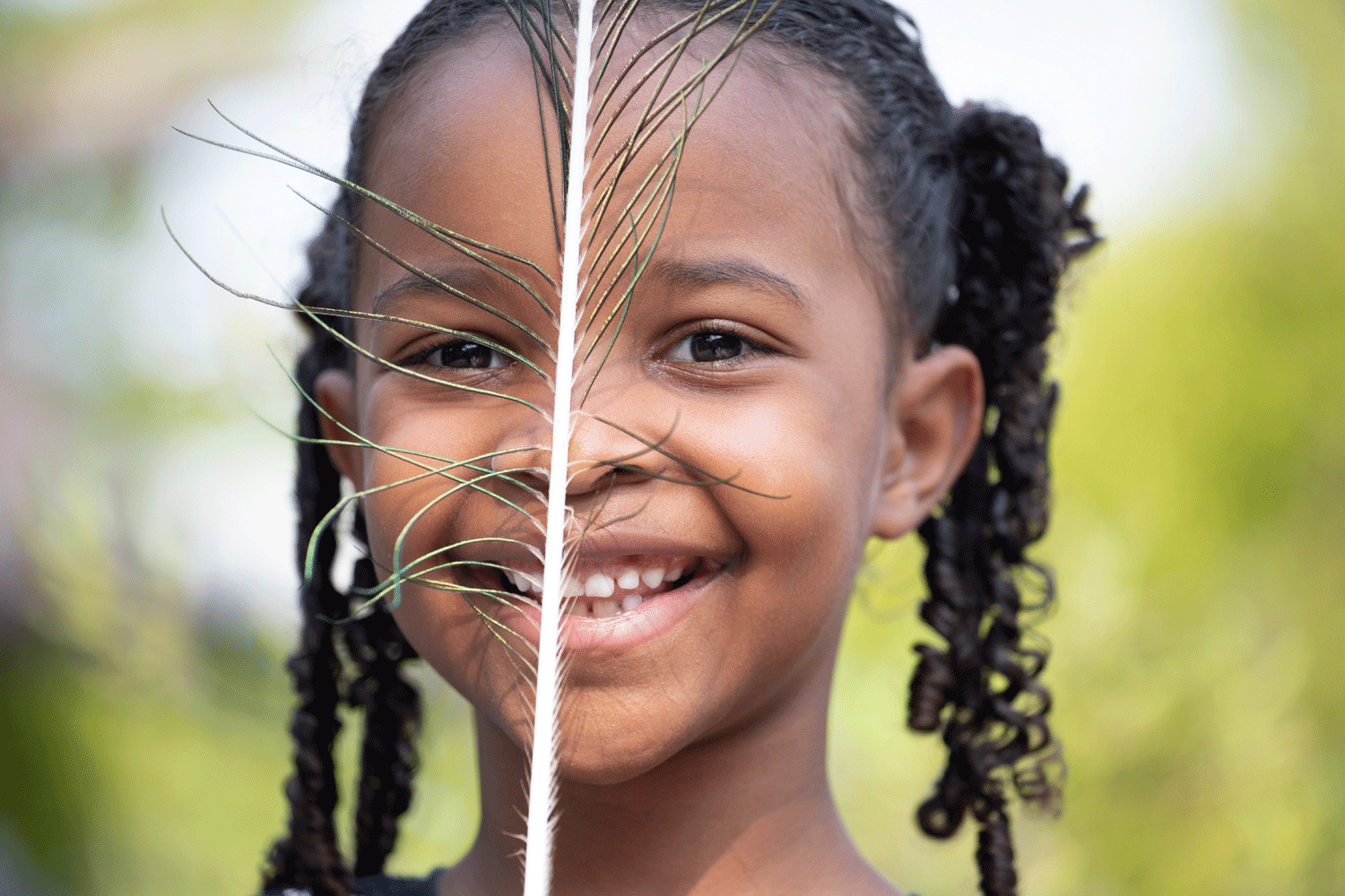 A smiling young girl holds a blue and green peacock feather in front of her face.