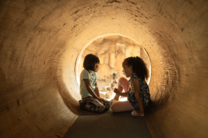Two young girls talk inside of a brown tunnel in the zoo.