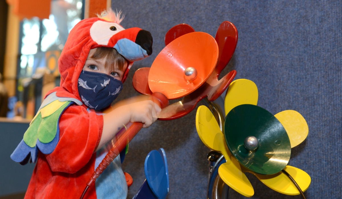 A young boy in a read parrot costume plays with colorful metal flowers that make music when tapped in the exhibit hall.