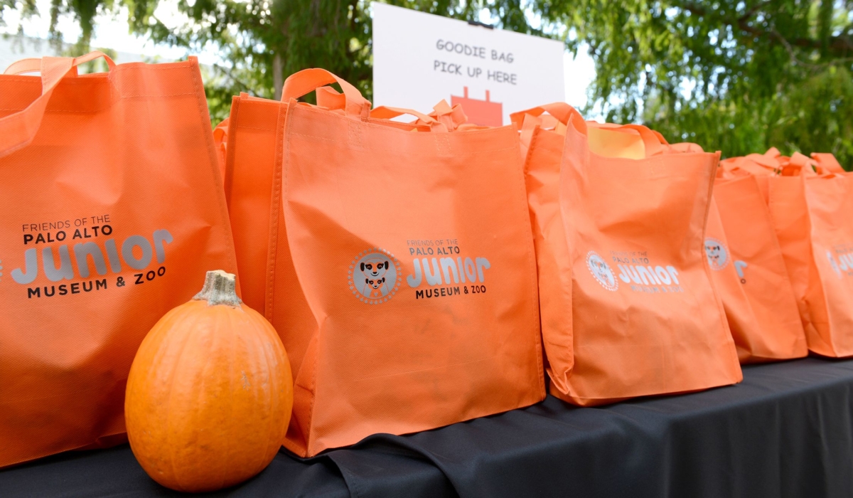 Orange goodie bags with the "Friends of the Palo Alto Junior Museum & Zoo" logo sit on a table with an orange pumpkin. The logo is black and gray and shows a meerkat mother and baby smiling.